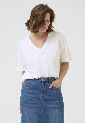 T-shirt blanc à col en V avec des clous décoratifs argentés sur les manches, associé à une jupe en jean ajustée. Tissu lisse et coupe décontractée.