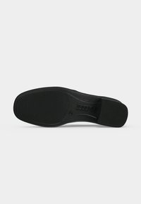 Semelle de chaussure noire avec grip en caoutchouc texturé au talon et à l’avant-pied, affichant le nom de la marque embossé dans la section centrale.
