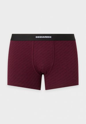 Un mannequin masculin se tient devant un fond clair uni, vêtu d’un boxer bordeaux à ceinture noire.