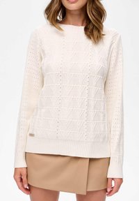 Kremowy sweter z dzianiny, z teksturowanym geometrycznym wzorem, z prążkowanymi mankietami i dołem. W zestawieniu z beżową spódnicą dla kontrastu.