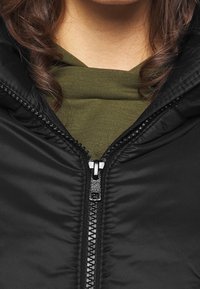 Detailní záběr na rozepnutou černou bundu přes olivově zelenou košili, ukazující detaily zipu a oblast límce se kudrnatými hnědými vlasy.