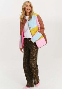 Žena s dlhými blond vlasmi, oblečená v pestrofarebnej prešívanej bunde, bielej košeli, nohaviciach s leopardím vzorom a ružových teniskách, stojí pred jednoduchým pozadím.