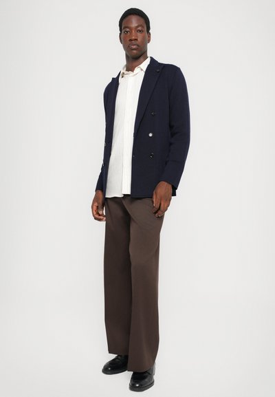 Navy double-breasted blazer, white striped shirt, brown wide-leg trousers, and black shoes. Model stands with hands at sides, neutral background.