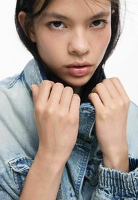 Geacă din denim cu guler bine croit, nuanță albastru deschis și textură uzată. Prim-plan cu mâini care apucă gulerul, cu piele netedă vizibilă.