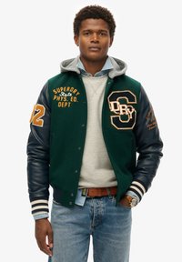 Grøn uld varsityjakke med marineblå læderærmer, prydet med orange broderi, en grå hætte og stribede ribkanter. Anbragt over en grå sweatshirt.