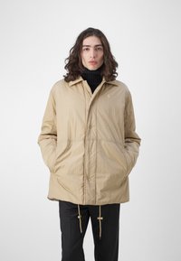 Khaki lichtgewicht jas met knoopsluiting, zijzakken en een trekkoord onderaan. Bevat een subtiele logo en een kraagontwerp.