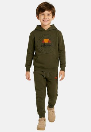 Jeune garçon souriant, portant un sweat à capuche olive de la marque Ellesse et un pantalon de jogging assorti, des baskets beiges, se tenant devant un fond blanc.