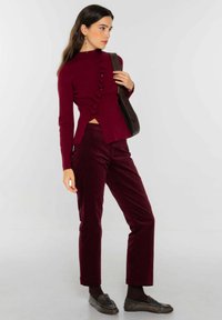 Top de manga larga color burdeos con detalle de volantes en el frente, combinado con pantalones de pana burdeos y zapatos marrones sin cordones, acompañado de un bolso de hombro.