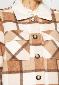 Brun och krämrutig skjortjacka tillverkad av mjukt, luddigt material. Har en krage, två framfickor och stora knappar.