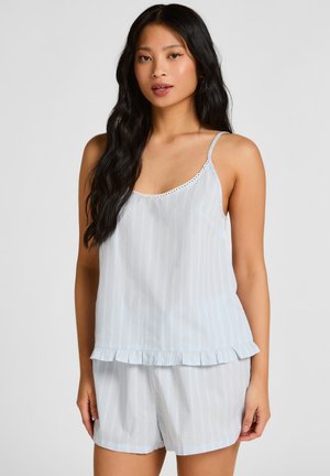 Jeune femme portant un haut sans manches rayé bleu clair et blanc et un short assorti, debout devant un fond uni.