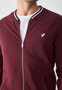 Bordo zip-up džemper s bijelim zatvaračem, bočnim džepovima i malim bijelim logom ptice na prsima. Ima prugasti rebrasti ovratnik.