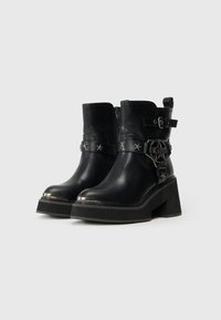 Botas de tornozelo em couro preto com saltos grossos, tachinhas em forma de estrela prateada, fivela decorativa e fecho lateral em fundo branco liso.