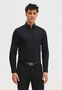 Czarna koszula z długim rękawem o dopasowanym kroju, z kołnierzykiem zapinanym na guziki oraz guzikami na przodzie. Noszona z czarnym paskiem, stylizowana z ciemnymi spodniami.