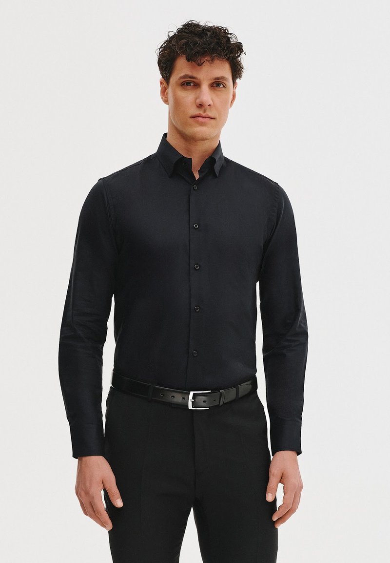 Czarna koszula z długim rękawem o dopasowanym kroju, z kołnierzykiem zapinanym na guziki oraz guzikami na przodzie. Noszona z czarnym paskiem, stylizowana z ciemnymi spodniami.