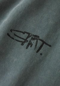 Black logo printed on a textured green fabric, featuring a continuous line design with a dot, emphasizing a smooth, soft surface.