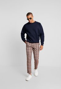 Mornarska dukserica s okruglim izrezom i rebrastim rukavima, uparena s smešnim kariranim hlačama i bijelim patikama. Na nosu su sunčane naočale.