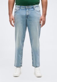 Άνδρας που φοράει ανοιχτό μπλε τζιν με ίσια γραμμή και λευκά αθλητικά παπούτσια, στέκεται μπροστά σε μια απλή ανοιχτή επιφάνεια υποβάθρου.