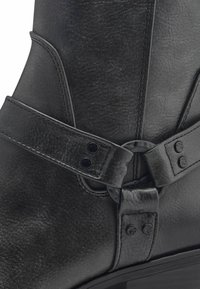 Bottine en cuir noir avec une large sangle dotée d'une boucle en métal circulaire, texture lisse et détails de couture visibles.