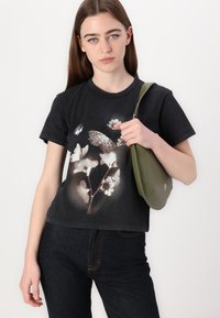 T-shirt nera corta con grafica di fiori e farfalle in tonalità tenui. Il modello tiene in mano una borsa verde con design curvo e chiusura con zip.