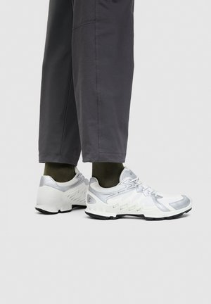 Witte en zilveren sportschoenen gedragen met donker groene sokken en losse donkergrijze broek, getoond vanaf halverwege de kuit naar beneden op een grijze achtergrond.