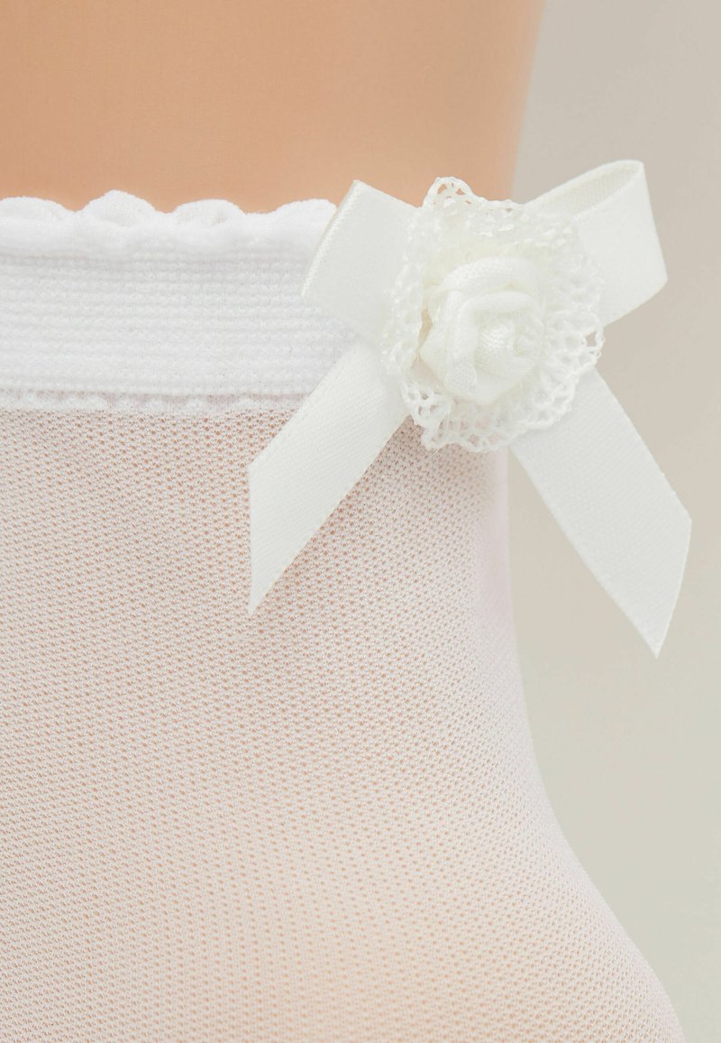 Collant bianchi con pizzo in cima realizzati in un tessuto trasparente e testurizzato. Presentano un piccolo fiore in tessuto e un nastro decorativo sul lato.