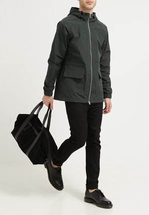 Homme marchant vêtu d'une veste à capuche noire, d'un pantalon noir et de chaussures noires, portant un grand sac de sport noir dans sa main gauche.