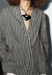 Personne portant un blazer rayé vertical noir et blanc avec un col en V profond et un grand pendentif noir sur un cordon.
