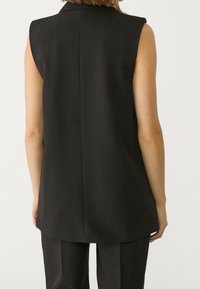 Musta hihaton liivi, jossa on jämäkkä kaulus, sileä kangas ja suora takasauma, yhdistettynä samanvärisiin mustiin housuihin.