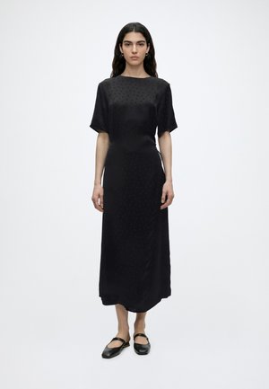 Kobieta z długimi ciemnymi włosami stoi zwrócona przodem, ubrana w czarną, krótką sukienkę sięgającą kostek i czarne, płaskie buty na jednolitym białym tle.