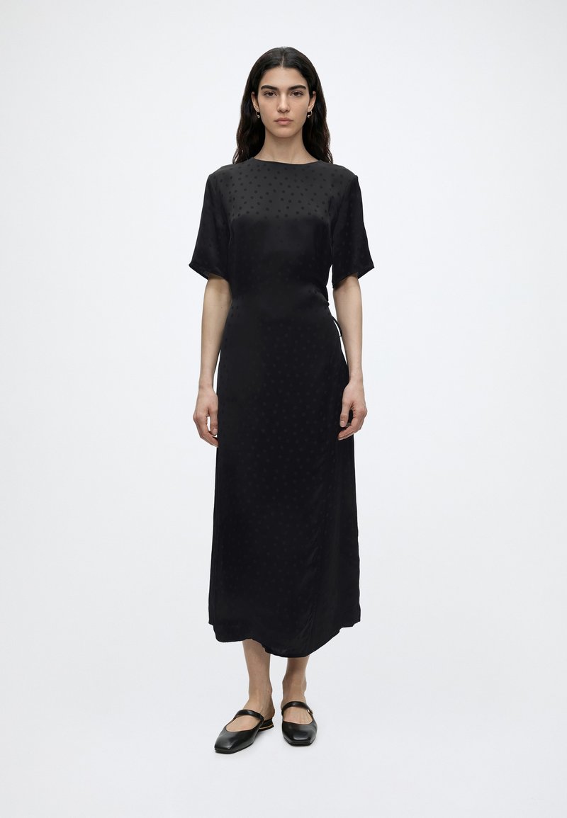 Γυναίκα με μακριά σκούρα μαλλιά στέκεται με το σώμα προς τα εμπρός φορώντας μαύρο φόρεμα με κοντά μανίκια μέχρι τους αστραγάλους και μαύρα επίπεδα παπούτσια, σε απλό λευκό φόντο.