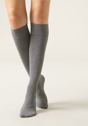 Piernas con calcetines largos hasta la rodilla, de color gris claro y sin costuras, sobre una superficie de tono beige claro, con los pies cruzados.