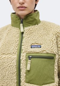 Άτομο που φοράει κρεμ φλις μπουφάν με πράσινες υφασμάτινες λεπτομέρειες και τσέπη στο στήθος με φερμουάρ, που έχει το λογότυπο Patagonia.