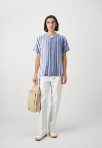 Short-sleeve blue and white striped button-up shirt, white trousers, beige backpack, and white sneakers with tan soles. Simple design, relaxed fit.