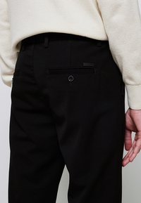 Personne portant un pantalon noir sur mesure avec une poche arrière boutonnée et un pull crème texturé, main détendue sur le côté.