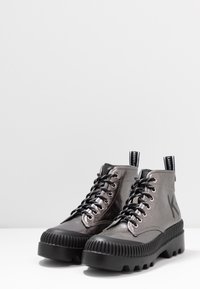 Un par de botas altas metálico grises con suelas gruesas negras, cordones negros y lengüetas de marca sobre una superficie blanca.