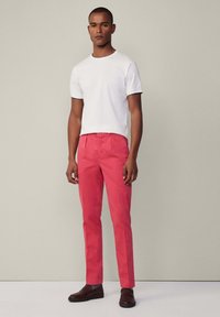 Rote Chino-Hosen mit geradem Schnitt, kombiniert mit einem weißen T-Shirt mit Rundhalsausschnitt. Das Modell trägt dunkelbraune Slipper. Einheitliche Farben, minimalistisches Design.