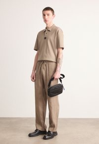 Beige poloshirt met korte mouwen; bijpassende beige elastische broek; zwarte schoudertas met rits; zwarte schoenen. Gladde stofstructuur.