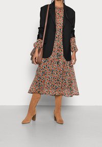 Vestido largo de flores con fondo negro, flores naranjas y blancas, diseño en capas. Blazer negro, bolso marrón, botas de tacón beige.