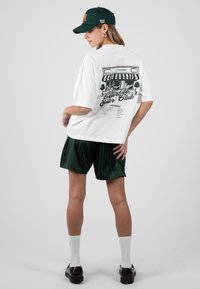 Femme portant un t-shirt blanc oversize avec un imprimé café noir dans le dos, un short vert foncé, des chaussettes blanches, des chaussures marron et une casquette de baseball vert foncé.