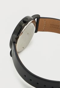 Montre avec bracelet en cuir noir, boîtier métallique rond, dos argenté, surface texturée et bouton texturé. Présente des éléments de design épurés.