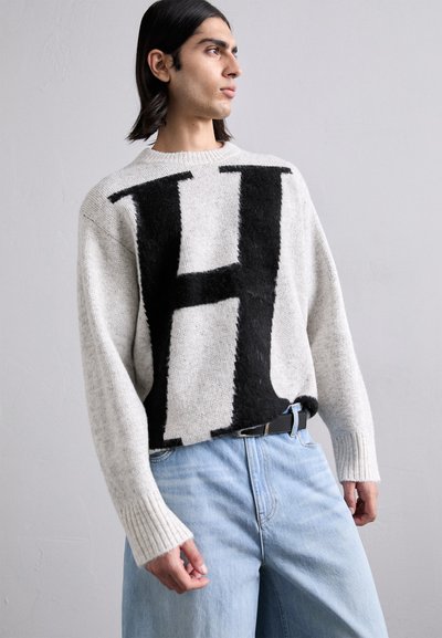 Levendula szürke, vastag kötésű pulóver, amelyen egy nagy fekete "H" minta található; bordázott mandzsetták és laza szabás, világoskék, széles szárú farmerral párosítva.