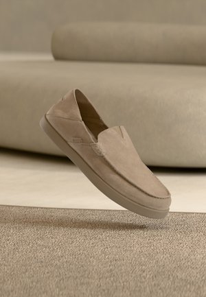 Beiger Wildleder-Slipper, der über einem strukturierten Teppich mit einem beigen Sofa im Hintergrund schwebt.