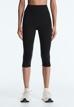 Osoba oblečená v čiernych legínach s vysokým pásom do kolien a v bielych športových topánkach s bielymi ponožkami, stojaca pred jednofarebným pozadím.