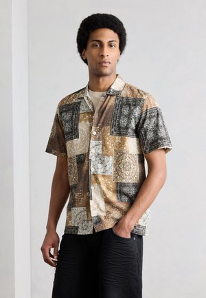 Muž s afrokuzlom, ktorý má na sebe krátky rukáv s práškovaným vzorom v zemských tónoch, stojí s jednou rukou v vrecku na jednotnom pozadí.