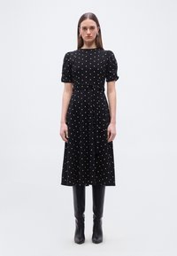 Γυναίκα που φοράει μαύρο midi φόρεμα με λευκές πουά, φουσκωτά μανίκια, ζώνη στη μέση και μαύρες μπότες μέχρι το γόνατο, στέκεται μπροστά σε ένα μονόχρωμο φόντο.