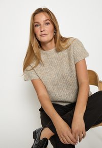 Bež lühikeste varrukatega kootud sviiter, millel on tekstureeritud muster ja ümar kaelus, koos mustade pükste ja läikivate mustade kingadega.