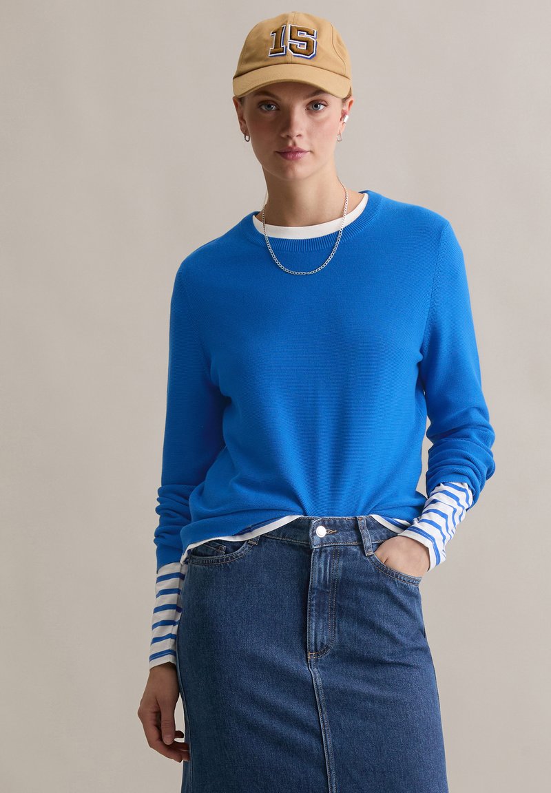 Blauwe geribbelde trui met lange mouwen en witte en blauwe strepen bij de manchetten, gedragen met een denim rok, geaccentueerd met een beige pet.