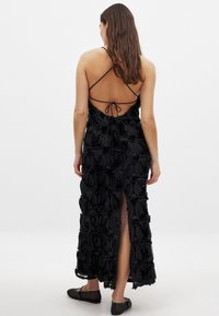 Vestido maxi negro con un diseño de rosas texturizadas, tirantes finos, espalda descubierta con cierre de lazo y una abertura lateral. Se llevan zapatos planos.