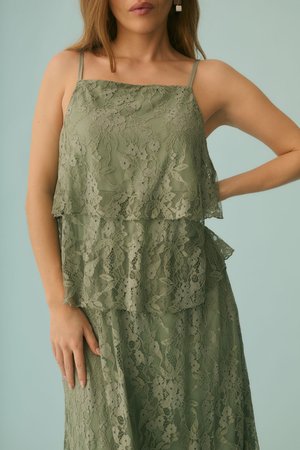 Femme en robe sans manches en dentelle vert olive avec un design en couches, debout avec une main sur la hanche devant un fond bleu clair uni.