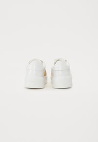 White sneakers with a textured tan panel on the side, smooth white heel, and raised white sole. Branding is embossed on the back.